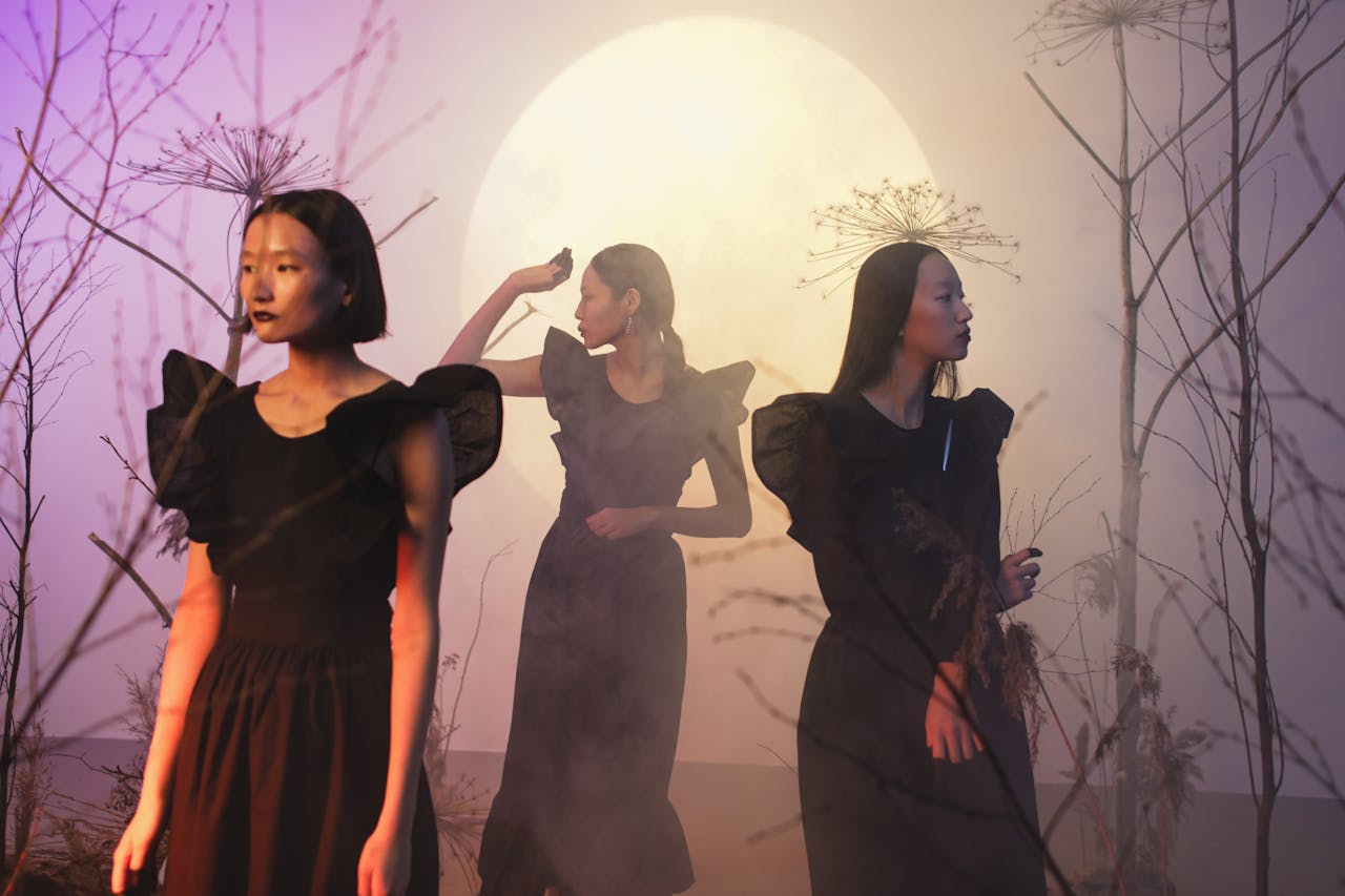 Three women in black dresses stand in a mysterious setting with moonlight and fog, creating a conceptual mood.